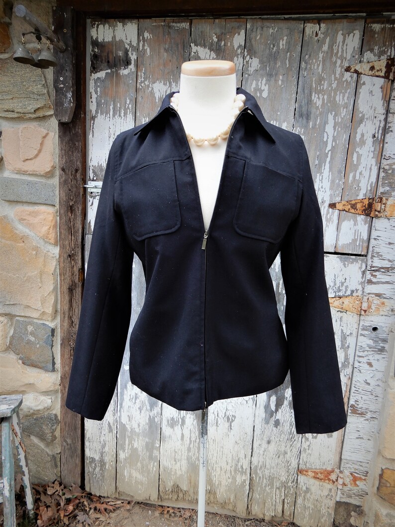 May include: A black zippered jacket with a collared neckline and two flap pockets on the front. The jacket is on a white mannequin and has a pearl necklace around the neck.