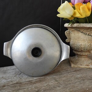 May include: A vintage aluminum pot lid with a black knob and side handles. The lid is silver and sits on a weathered wooden surface. A decorative urn with colorful flowers is in the background.