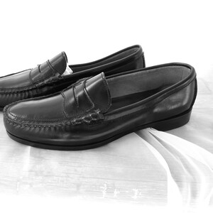 May include: A pair of black leather loafers with a penny strap detail. The loafers are on a white fabric surface.