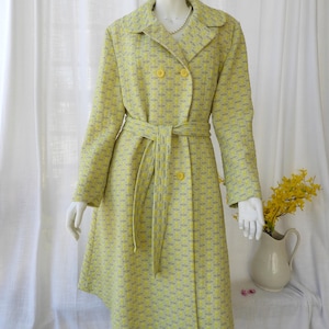 May include: A vintage-style coat in a light yellow and gray geometric pattern. The coat features a double-breasted design with large yellow buttons, a matching belt, and a classic collar. The coat is displayed on a mannequin.