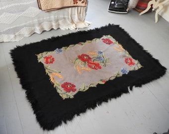 Puede incluir: Una alfombra vintage con un borde negro con flecos y un centro gris con un diseño floral con amapolas rojas, flores azules y trigo amarillo.
