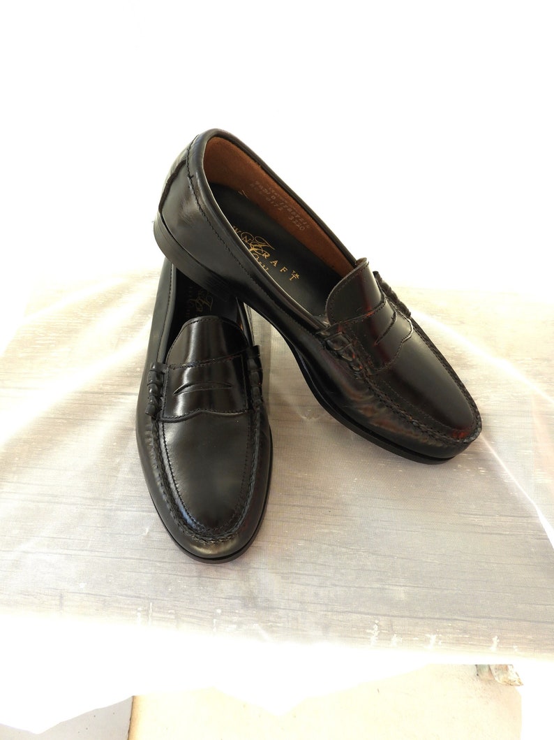 May include: A pair of black leather loafers with a penny strap detail. The loafers have a brown leather lining and a gold embossed logo on the insole.
