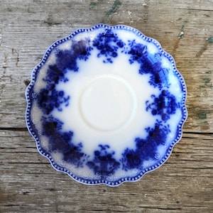 May include: A white and blue saucer with a scalloped edge. The saucer features a floral design in shades of blue around the rim and a white center. The rim has a delicate dotted pattern. The saucer is likely vintage or antique.