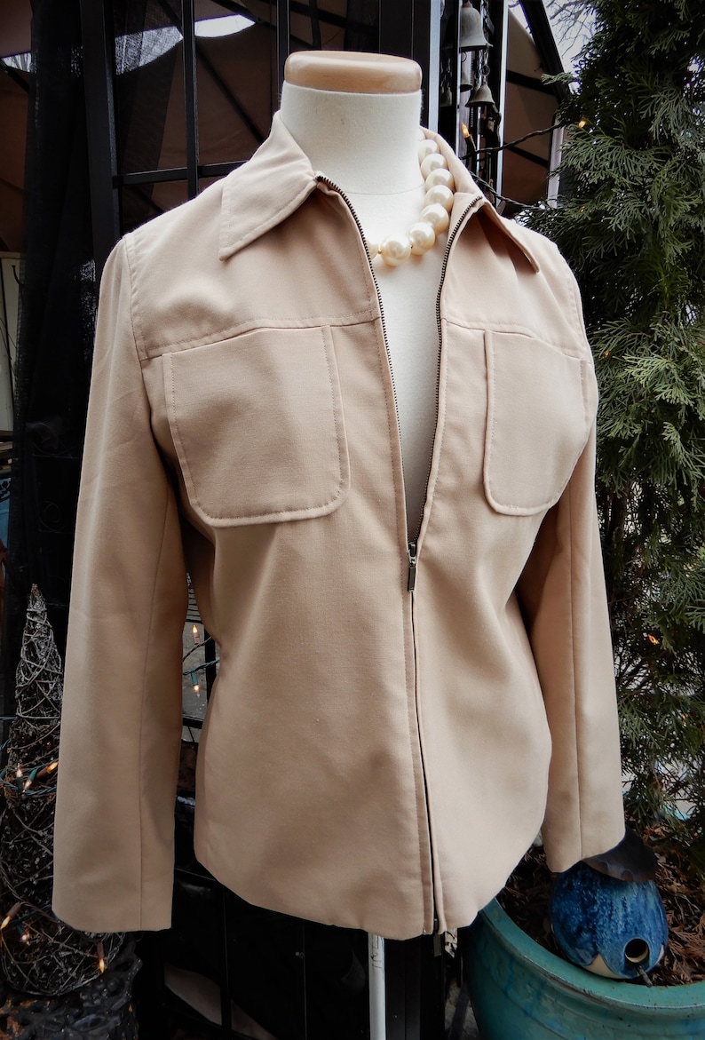 May include: A beige zip-up jacket with a collar and two large pockets on the front. The jacket is on a mannequin wearing a pearl necklace.
