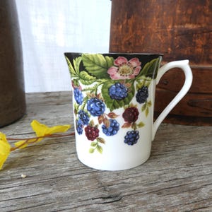 May include: White ceramic mug with a handle, decorated with a pattern of blackberries, pink flowers, and green leaves. The rim of the mug is black. The mug is on a wooden surface.