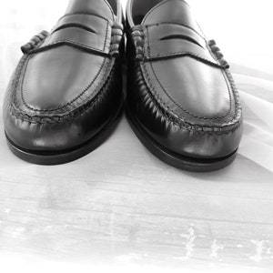May include: A pair of black leather loafers with a stitched detail around the edge. The loafers are on a white fabric background.