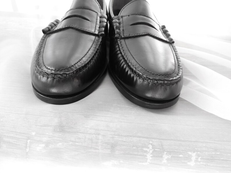 May include: A pair of black leather loafers with a stitched detail around the edge. The loafers are on a white fabric background.