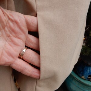 May include: A beige blazer with a single pocket on the left side. The pocket is open and a hand is reaching into it. The hand is wearing a gold ring.