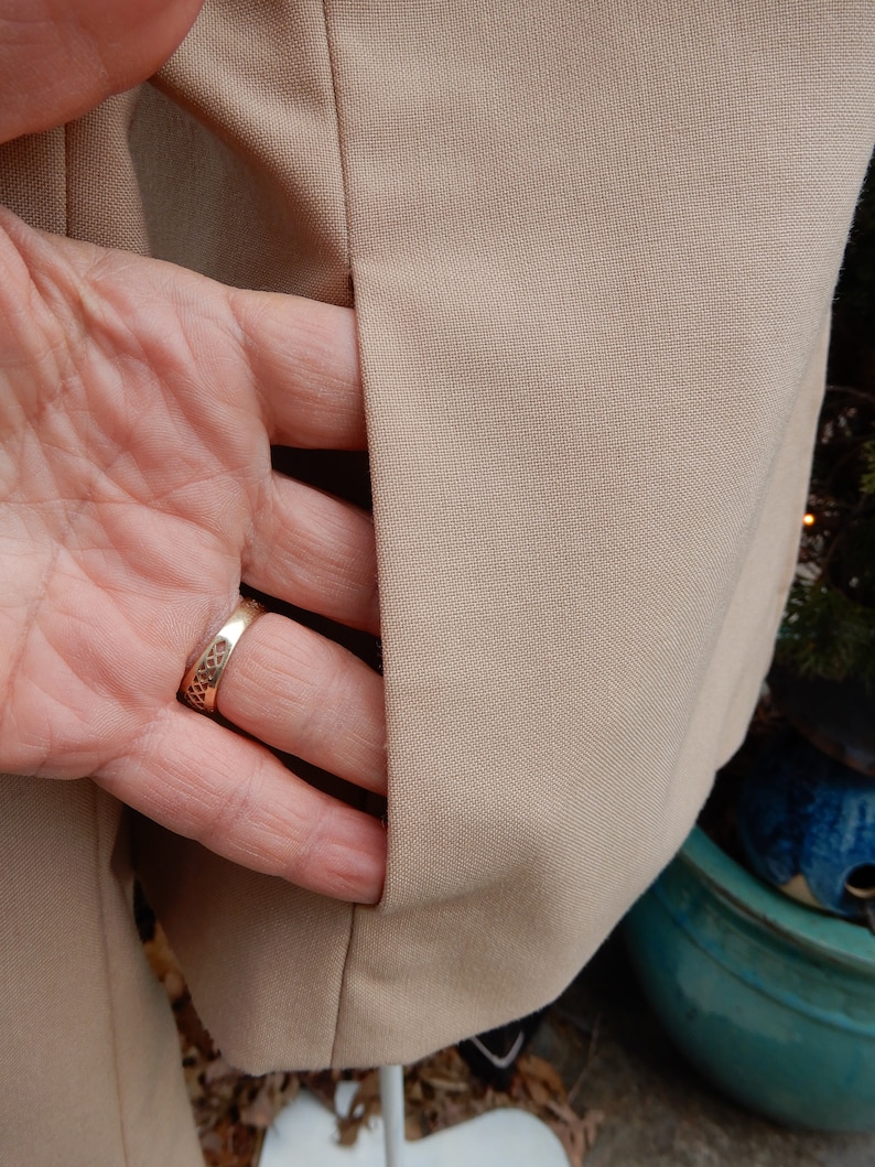 May include: A beige blazer with a single pocket on the left side. The pocket is open and a hand is reaching into it. The hand is wearing a gold ring.