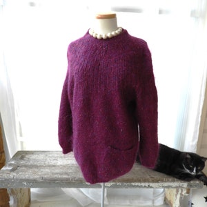 May include: A deep purple knitted sweater with a small pocket on the front. The sweater is on a mannequin wearing a pearl necklace.