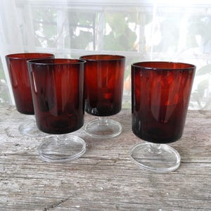 May include: Four clear glass stemmed drinking glasses with a dark red, almost burgundy, hue. The glasses are on a rustic wooden surface.