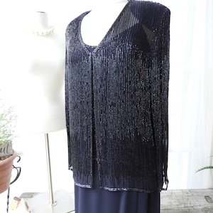 May include: A black beaded cardigan with a V-neckline and long sleeves. The cardigan is made of small, black beads that are strung together to create a fringe effect. The cardigan is being displayed on a white mannequin.