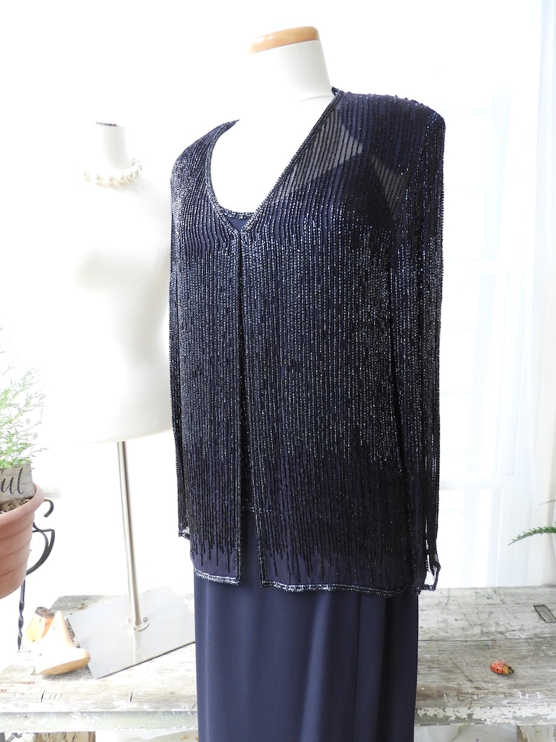 May include: A black beaded cardigan with a V-neckline and long sleeves. The cardigan is made of small, black beads that are strung together to create a fringe effect. The cardigan is being displayed on a white mannequin.