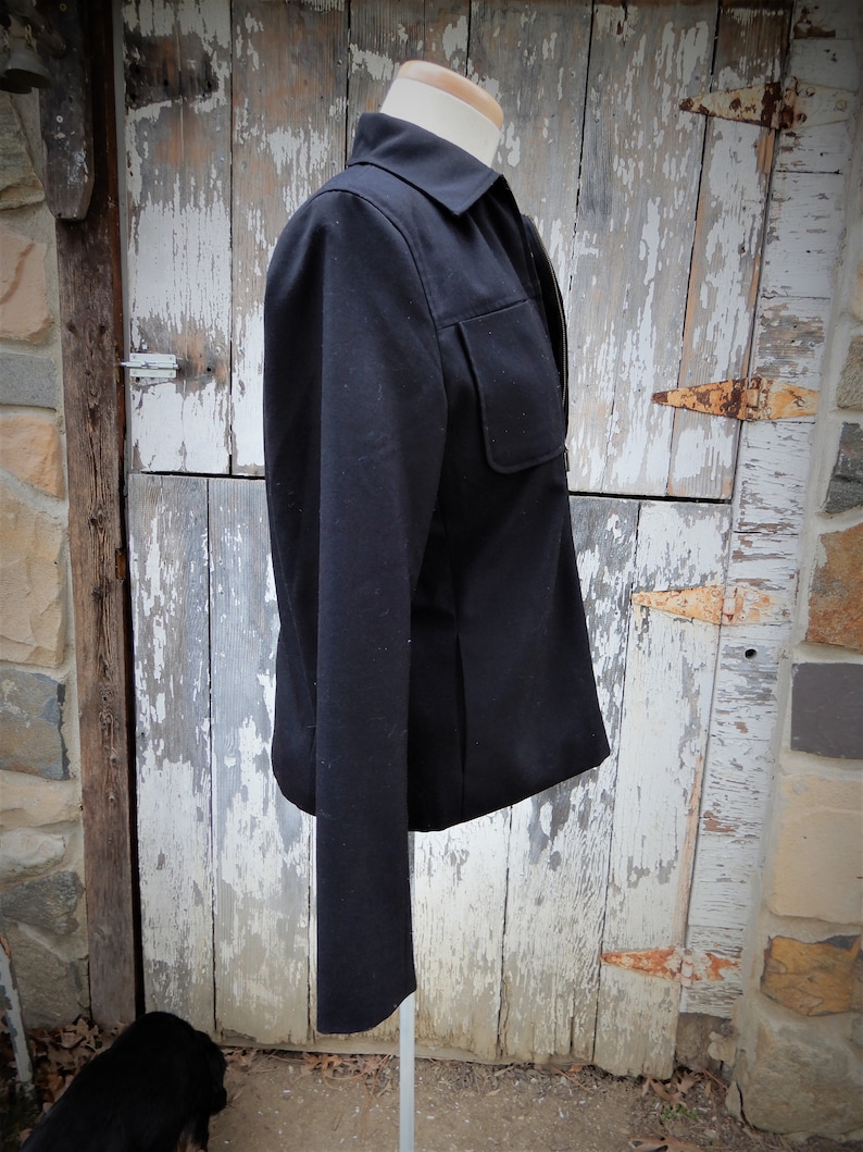 May include: A black blazer with a zipper closure and a single pocket on the left side. The blazer is on a white mannequin and has long sleeves.