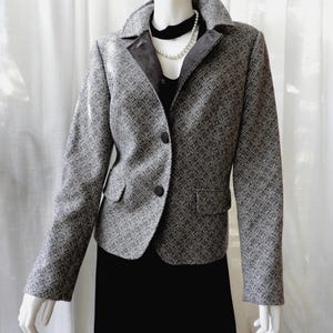 May include: A grey and white patterned blazer featuring a notched collar, long sleeves, and two front buttons. The jacket has dark grey lapel facings and two flap pockets at the waist, worn over a black top with a white pearl necklace.
