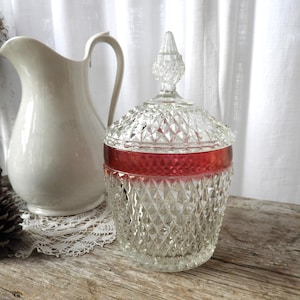 Puede incluir: Un cuenco de cristal transparente con una banda roja y un patrón de diamantes. El cuenco tiene una tapa a juego con un remate decorativo. Una jarra blanca está en el fondo.