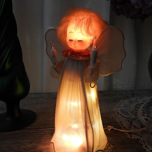 May include: A lighted angel figurine with orange hair, closed eyes, and long eyelashes. The figure wears a white, translucent, floor-length gown illuminated from within by small lights. It holds a small white candle in each hand with wings.