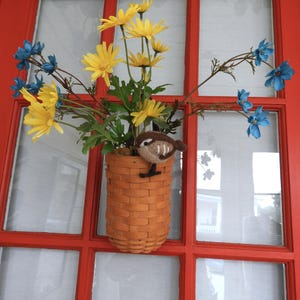 May include: A woven light brown basket hanging on a red door, filled with yellow daisy-like flowers and blue blossoms. A small, brown, needle-felted bird figure with white wing stripes is perched on the rim of the basket among the green stems.