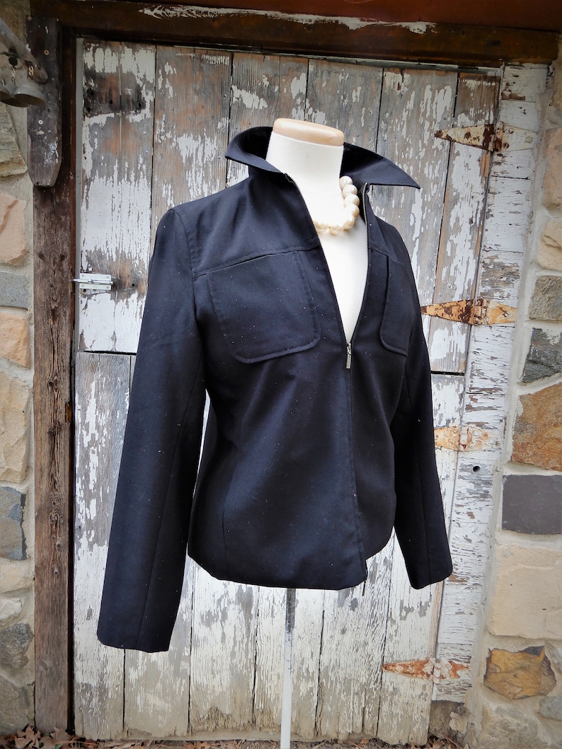 May include: A black zippered jacket with a stand-up collar and a single pocket on the left side. The jacket is on a white mannequin against a weathered wooden background.