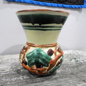 May include: A ceramic vase with a wide mouth and a narrow base. The vase is decorated with green, brown, and orange paint. The vase has a stylized design of trees and branches.