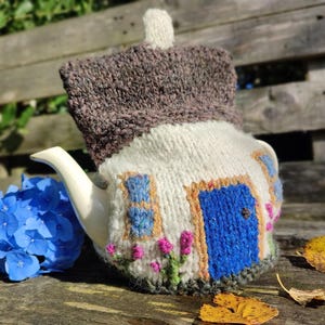 Può includere: Una teiera in ceramica bianca con una coperta di lana a forma di cottage. La coperta presenta un tetto marrone, una porta blu e finestre con accenti floreali rosa e verdi. La teiera è appoggiata su una superficie di legno con fiori blu e foglie cadute.