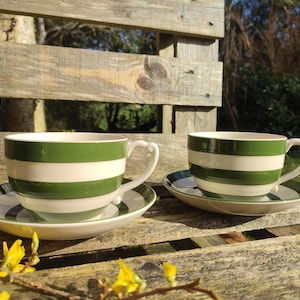 Puede incluir: Dos tazas de té y platillos de cerámica con rayas horizontales verdes y blancas. Las tazas tienen un asa curva y están sobre una superficie de madera. Los platillos también tienen rayas verdes y blancas.