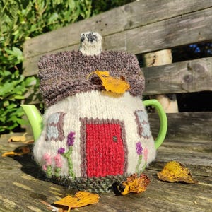 Könnte beinhalten: Eine skurrile, handgestrickte Teekannenhaube in Form eines Häuschens. Die Haube hat ein braunes Dach, eine rote Tür und gestickte Blumen. Die Teekanne hat einen grünen Griff und Ausguss. Gelbe Blätter liegen auf der Holzoberfläche.