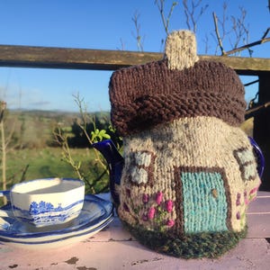 May include: A whimsical, hand-knitted teapot cozy shaped like a cottage, with a brown roof and a blue door. A matching blue and white teacup and saucer sit nearby. The cozy is beige with pink and green floral accents.