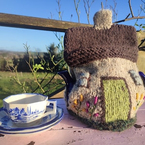 May include: A charming, hand-knitted tea cosy in the shape of a cottage, featuring a brown roof and green door. A matching teacup and saucer with a blue and white landscape design are on a pink table, creating a cosy tea setting.