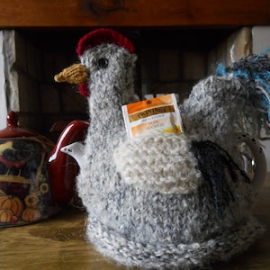 May include: A handmade gray chicken tea cozy with a red comb and yellow beak. A tea bag is tucked into the cozy. A decorative teapot with a fall harvest design is in the background.