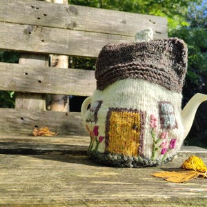 Könnte beinhalten: Eine weiße Keramik-Teekanne mit einem gestrickten Überzug in Form eines Häuschens. Der Überzug hat ein braunes Dach, eine gelbe Tür und rosa Blumenakzente. Die Teekanne steht auf einer Holzbank mit Herbstblättern.