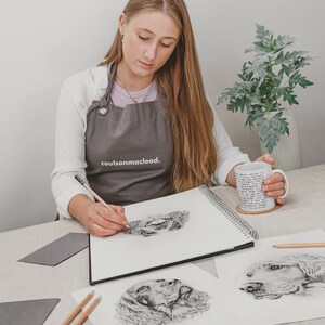 May include: An artist sketching a detailed pencil drawing of a dog's eye in a spiral-bound sketchbook. The artist wears a gray apron with the text "coulsonmacloed." A mug with text is held in the artist's hand. Additional dog portraits are on the table.
