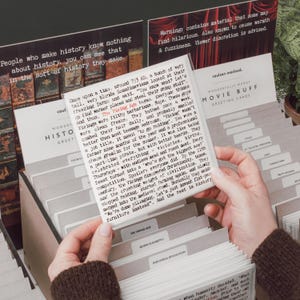 May include: A person holds a card from a card catalog labeled "Wonderfully Wordy Movie Buff Greeting Cards." The card features text about the Viking Age. Other cards are visible, including "Queen Elizabeth" and "Industrial Revolution."