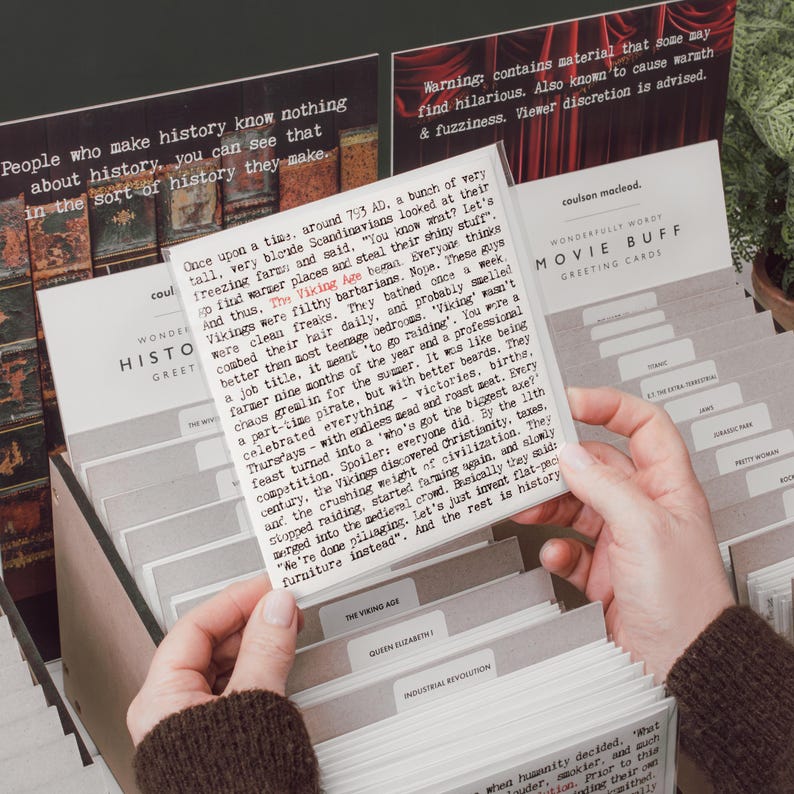 May include: A person holds a card from a card catalog labeled "Wonderfully Wordy Movie Buff Greeting Cards." The card features text about the Viking Age. Other cards are visible, including "Queen Elizabeth" and "Industrial Revolution."