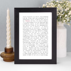 May include: A framed print with black frame features the text "Great theatre is about challenging how we think..." in a black font. A white candle and a vase of white flowers are on either side of the framed print.