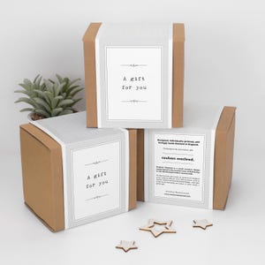 Może przedstawiać: Trzy brązowe kartonowe pudełka na prezenty z białymi etykietami z napisem "A gift for you". Pudełka są ułożone w stos, a w tle znajduje się mała roślina i drewniane dekoracje w kształcie gwiazd.