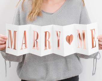 以下が含まれることがあります： メタリックなローズゴールドの箔押しで「MARRY ME」と書かれた、アコーディオン状に折りたたまれた白いカード。文字の間にはローズゴールドのハートのシンボルが配置されており、白い背景で視認性が高められています。