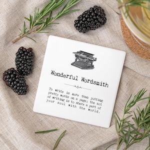 May include: A white square coaster featuring the words "Wonderful Wordsmith" and a vintage typewriter illustration. Fresh blackberries and rosemary sprigs are arranged around it.