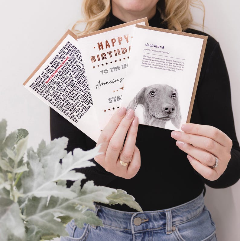 Pu&ograve; includere: Tre biglietti di auguri sono tenuti davanti a una persona. I biglietti hanno testo e immagini. Un biglietto dice "Happy Birthday" con stelle. Un altro biglietto ha un'illustrazione di bassotto. Il terzo biglietto ha un elenco di testo.