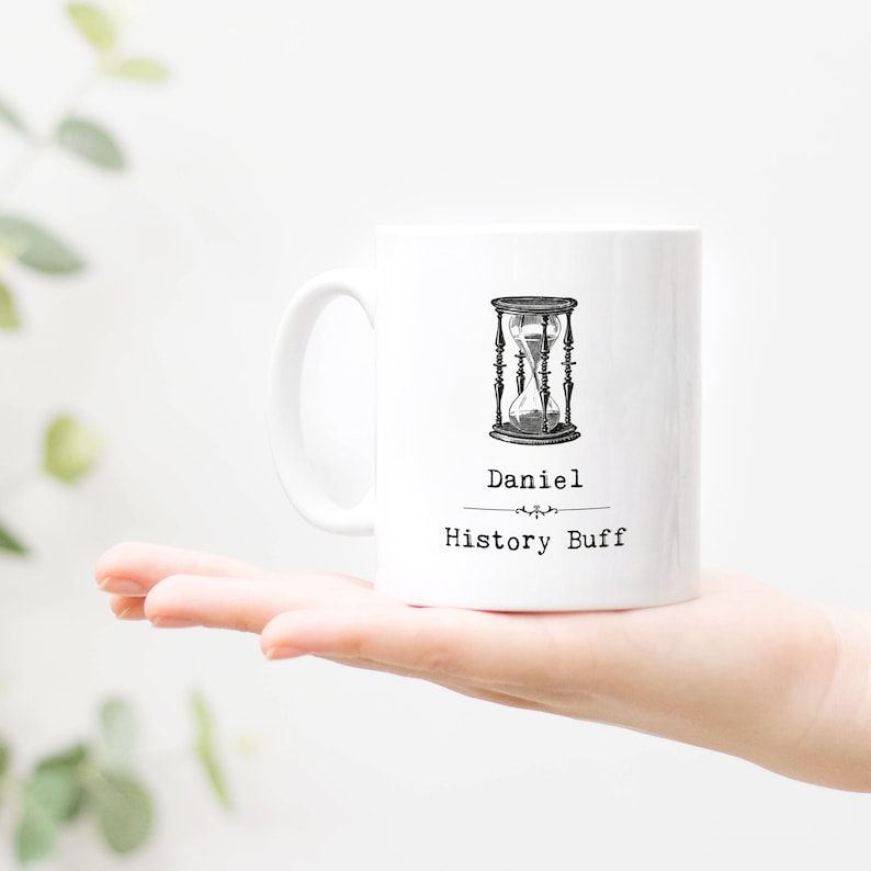 Może przedstawiać: Biały ceramiczny kubek z czarno-białą ilustracją klepsydry. Na kubku widnieje imię "Daniel" i napis "History Buff". Kubek jest trzymany w dłoni osoby.