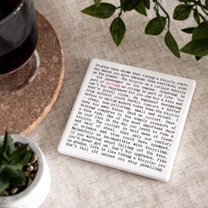 May include: A square, white ceramic coaster with black text about cycling. The coaster is on a neutral-toned surface, next to a glass of red wine and a small potted plant. The text includes the word "Cycling".