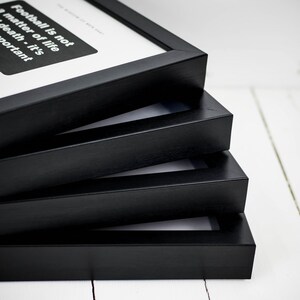 May include: A stack of four black picture frames. The top frame displays text that reads "Football is not a matter of life or death - it's important" in white font on a black background. The frames are stacked on a white wooden surface.