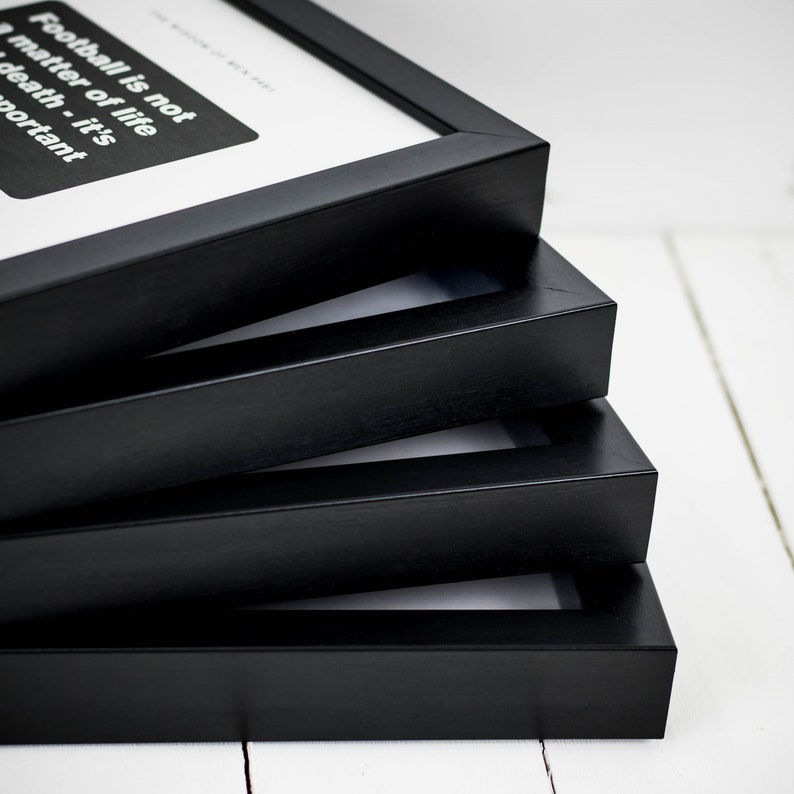 May include: A stack of four black picture frames. The top frame displays text that reads "Football is not a matter of life or death - it's important" in white font on a black background. The frames are stacked on a white wooden surface.
