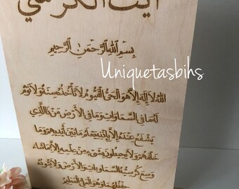 May include: A light-colored wooden plaque with Arabic calligraphy, featuring the text "Ayat al-Kursi." The plaque is mounted on a rectangular base. The text "Uniquetasbihs" is also visible on the plaque.