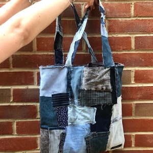 May include: A blue patchwork tote bag made from recycled denim. The bag has a large rectangular shape with two long straps. The bag is made from different shades of blue denim and has a variety of textures.