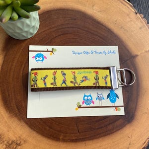 May include: A yellow and brown keychain with a repeating pattern of cartoon animals. The keychain has a silver metal clasp. The card behind the keychain reads "Unique Gifts & Treats by Shelia" and features owl illustrations.