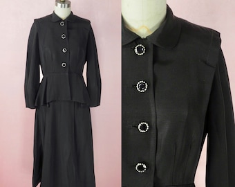 May include: A black vintage dress suit with a fitted jacket and a skirt. The jacket has a peter pan collar and features a row of black buttons with rhinestones. The skirt is knee-length and has a slight A-line shape.