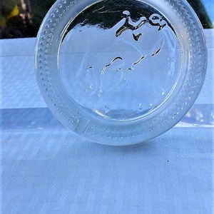 May include: A clear glass ashtray with a raised design of an elephant with its trunk raised. The ashtray has a beaded edge.