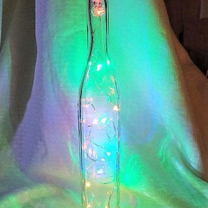 May include: A clear glass bottle with a cork stopper filled with white fairy lights. The bottle is illuminated with a green and blue glow.