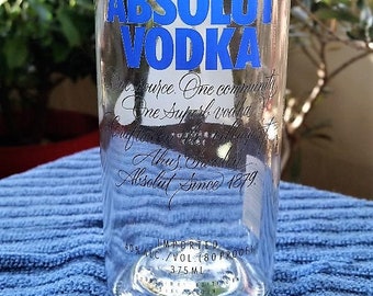 Könnte beinhalten: Eine durchsichtige Glasflasche mit einem blau-weißen Etikett, auf dem "ABSOLUT VODKA" steht. Das Etikett enthält auch den Text "One source. One community. One superb vodka." und "Absolut Since 1879." Die Flasche steht auf einer blauen Oberfläche.